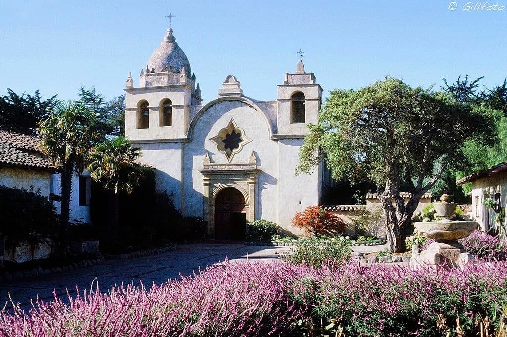 Exterior of Spanish style church with trees and flowering garden in the foreground.