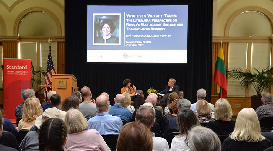 People are sitting in a large conference room listening to a woman and a man who are sitting on armchairs on a stage.